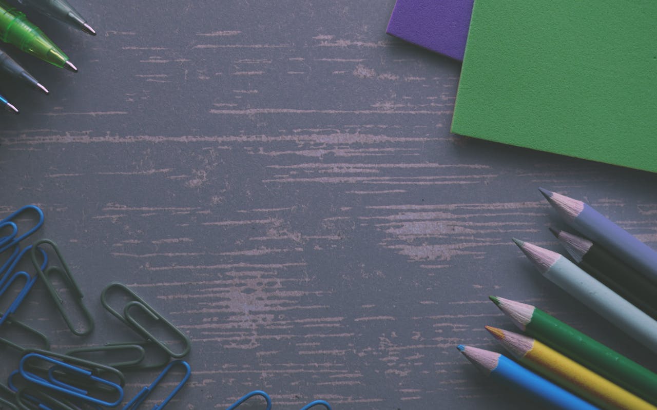 A flat lay of colored pencils, paper clips, and paper on a wooden desk.